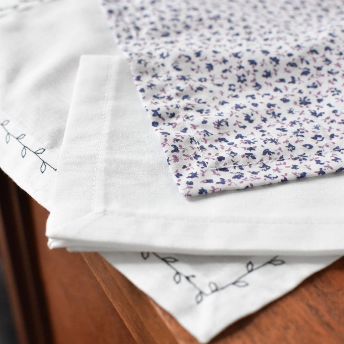 Folded white DIY handkerchiefs feature delicate floral embroidery on the edges with one a blue and purple small floral pattern.
