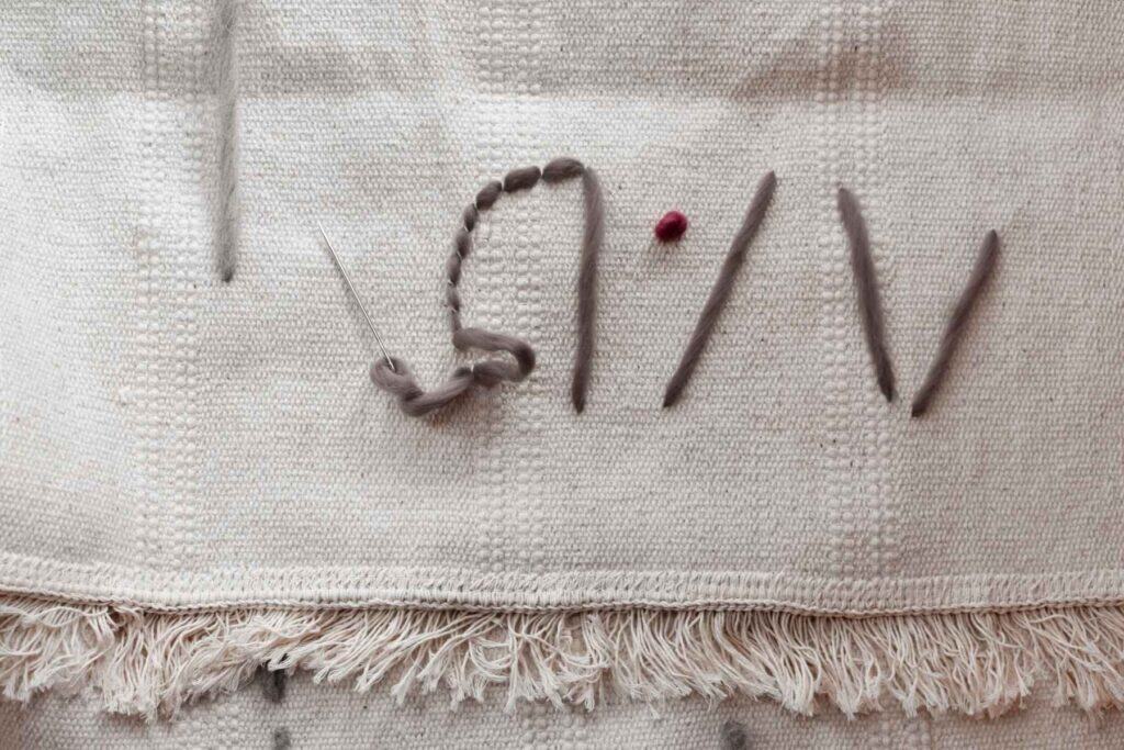 A white woven fabric wall hanging with a fringe at the bottom displays gray stitches and a single red dot. The design forms abstract lines and shapes on the textured surface, creating a tapestry-style look.