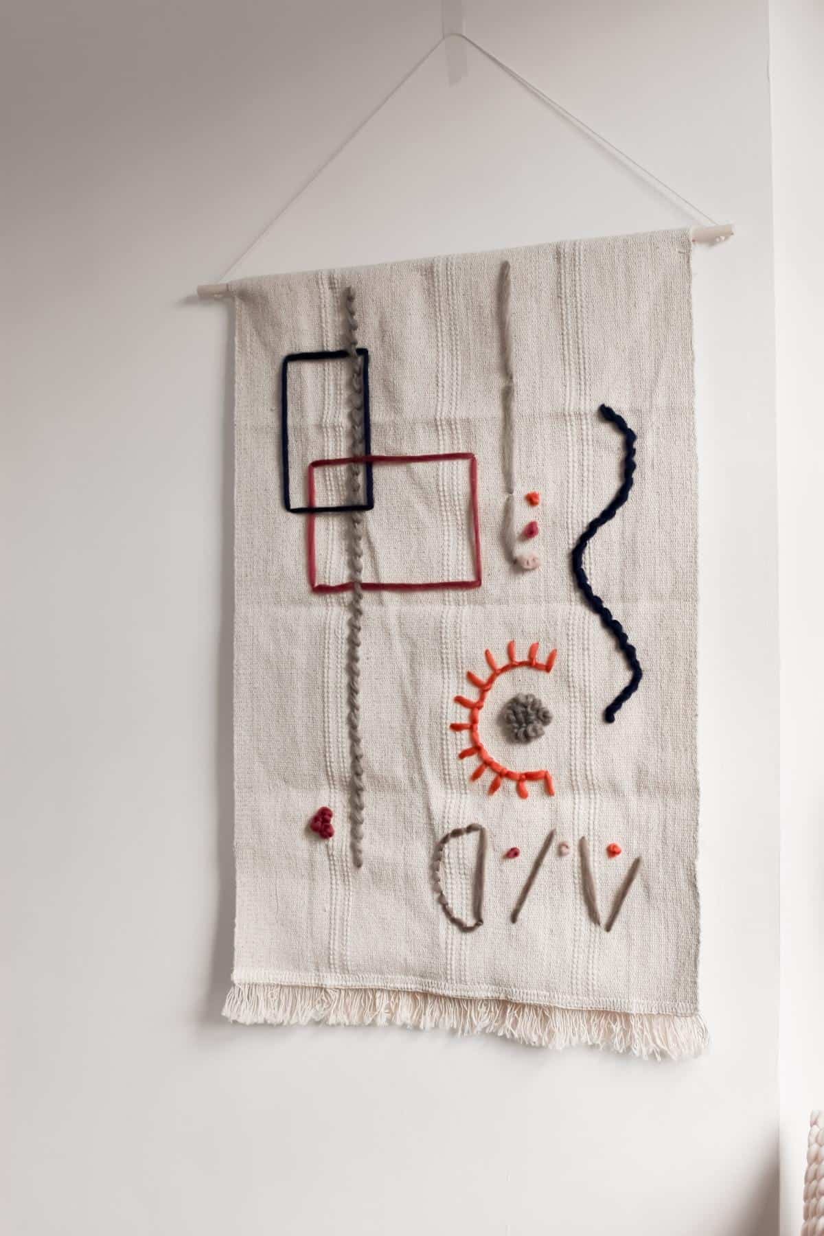 A textured tapestry features abstract designs, showcasing red and navy rectangles, a sun pattern, wavy lines, and shapes in gray, red, and black. This quick addition to decor hangs on a white wall with a simple string.