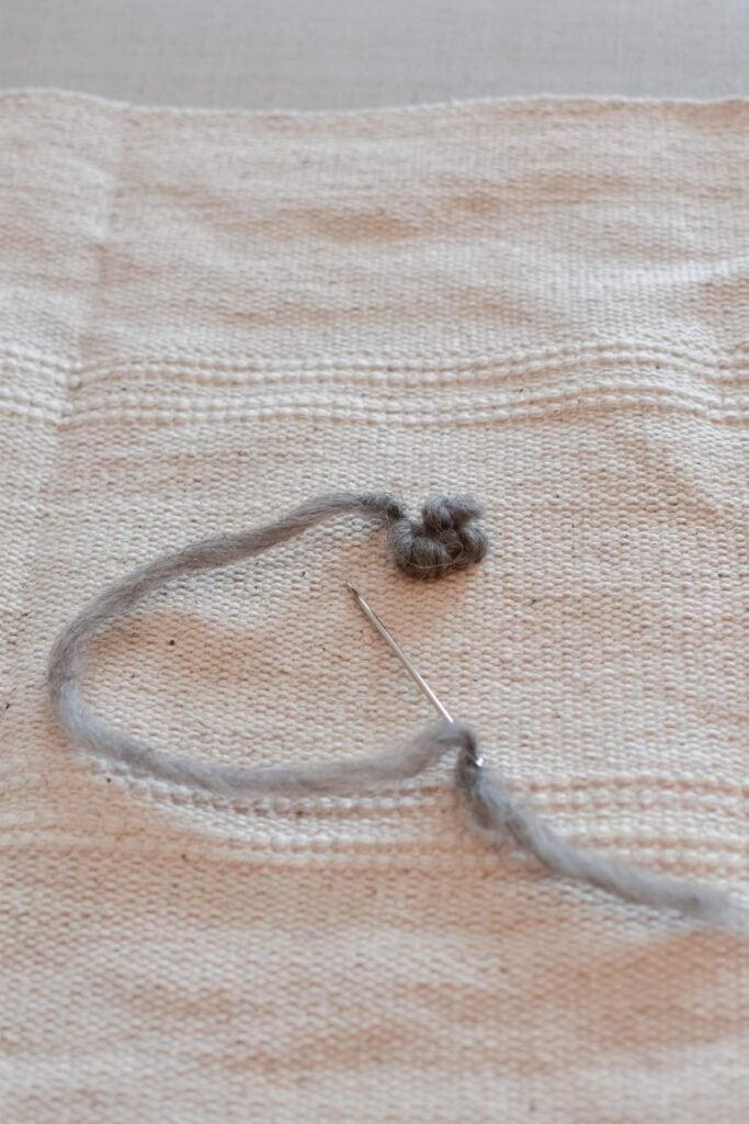 A needle and gray thread are partially sewn into a textured beige fabric, forming a small looped design. 