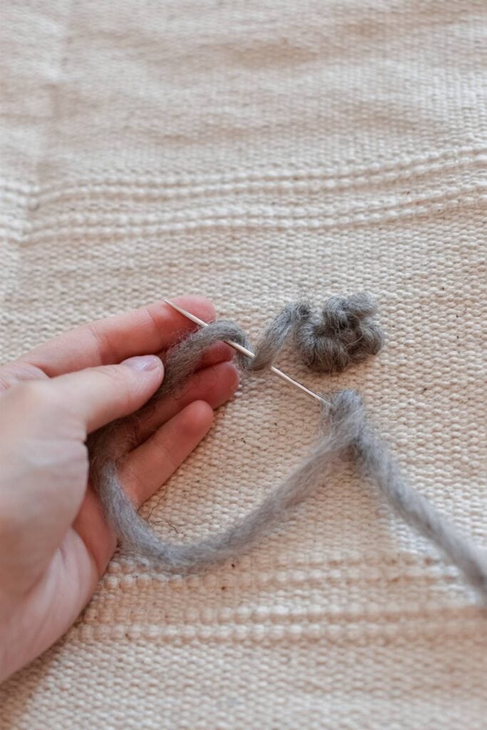 A hand skillfully maneuvers a needle and gray yarn, demonstrating a quick sewing technique on a textured beige fabric, perfect for creating an elegant tapestry or wall hanging.