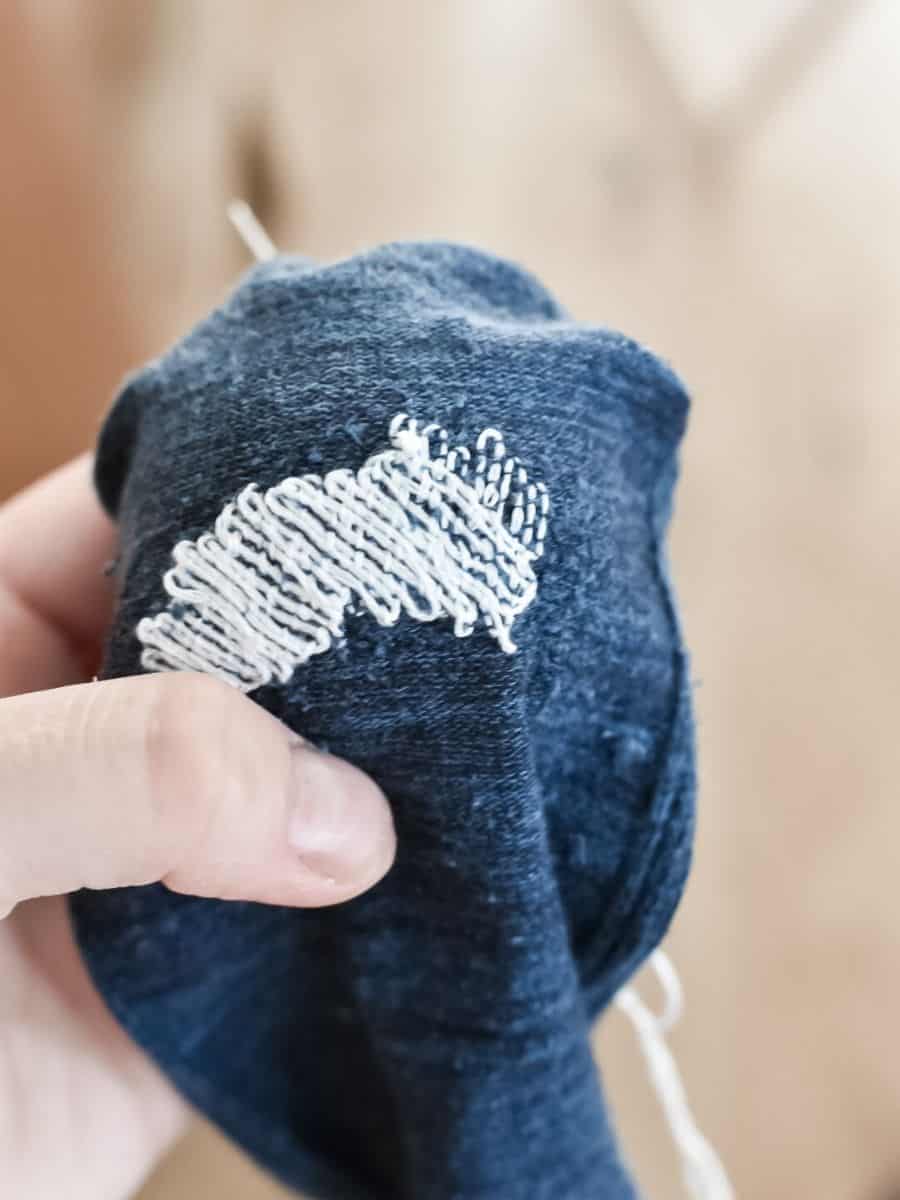 A hand holding a dark blue sock with white darning stitches neatly reinforcing a worn-out area.