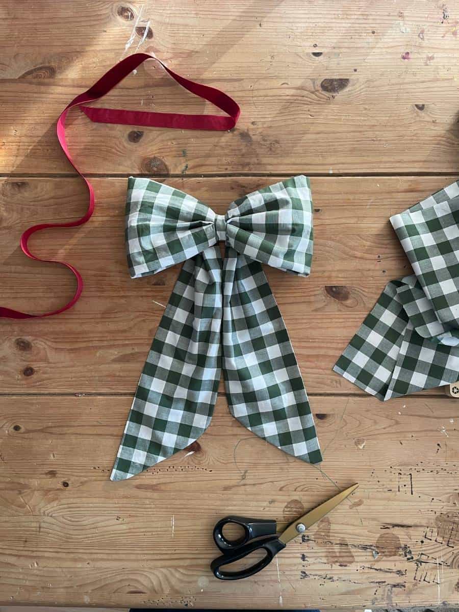 A green and white gingham fabric bow on a wooden table, with a red ribbon, fabric scraps, and sewing scissors nearby—preparing materials for a handmade bow.