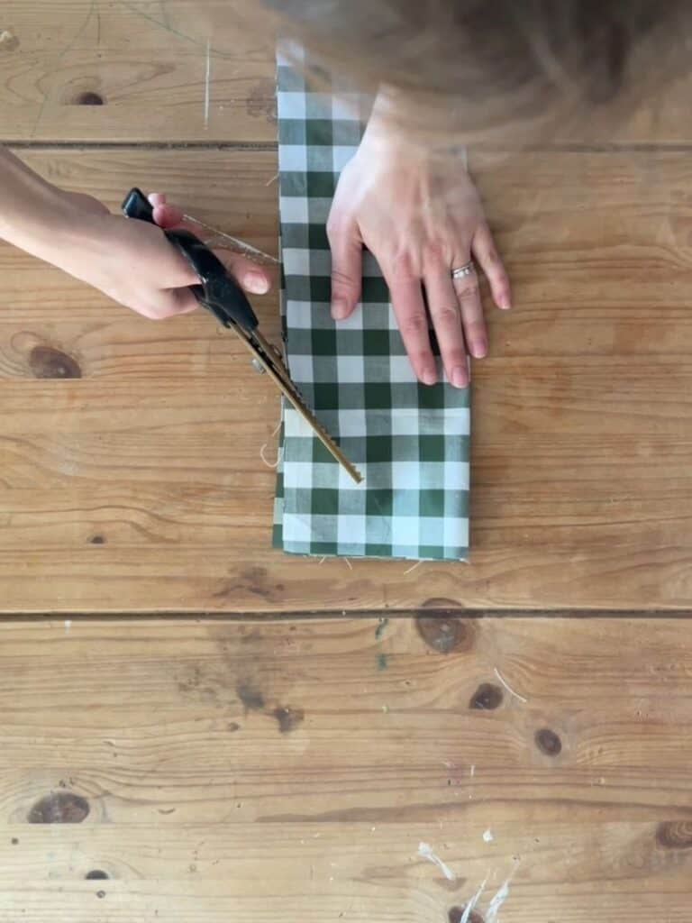 A person uses pinking shears to trim the edge of folded gingham fabric, ensuring a neat and fray-resistant finish for the bow tails.
