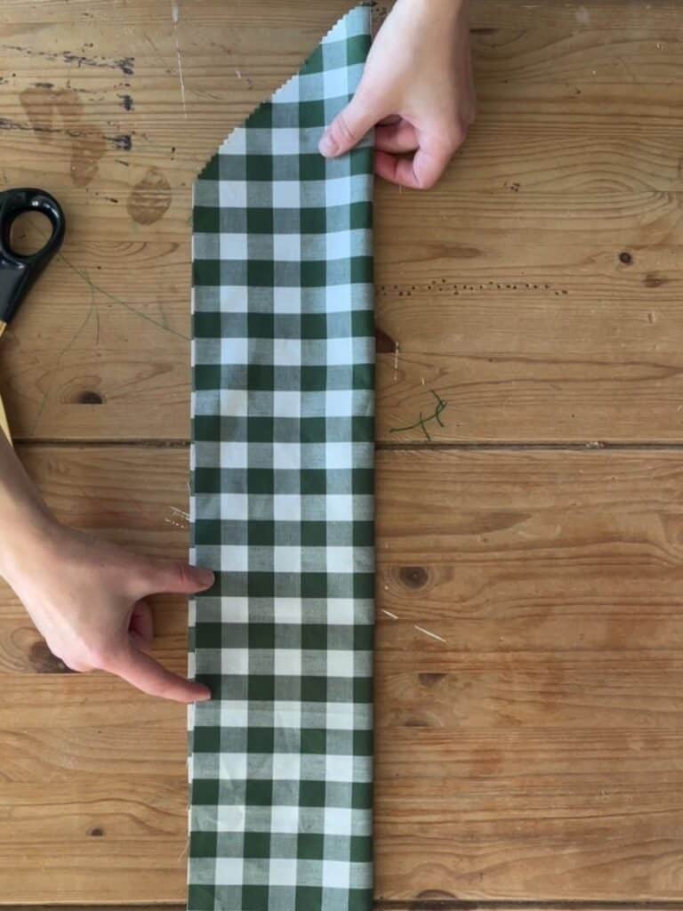 Hands unfolding a long strip of gingham fabric with one pointed end, showing the shaping process for the bow tails.