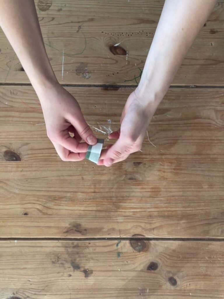 A small strip of gingham fabric is carefully folded and pinched between two hands, preparing the center knot piece for the bow.