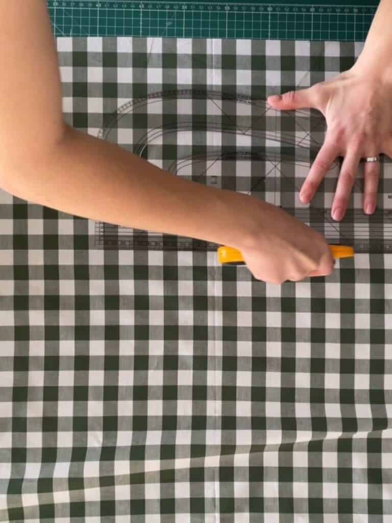 Hands measuring and marking the gingham fabric using a rotary cutter and ruler, carefully cutting the pieces for the bow.