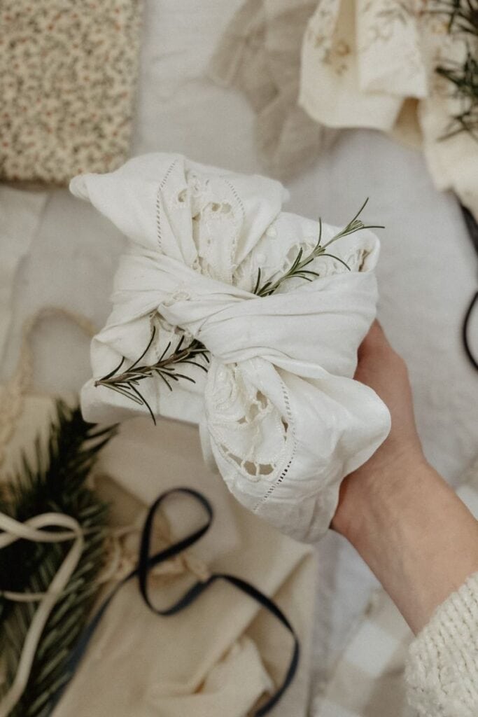 A beautifully wrapped gift in vintage white embroidered fabric, tied with a knot and adorned with a sprig of rosemary.