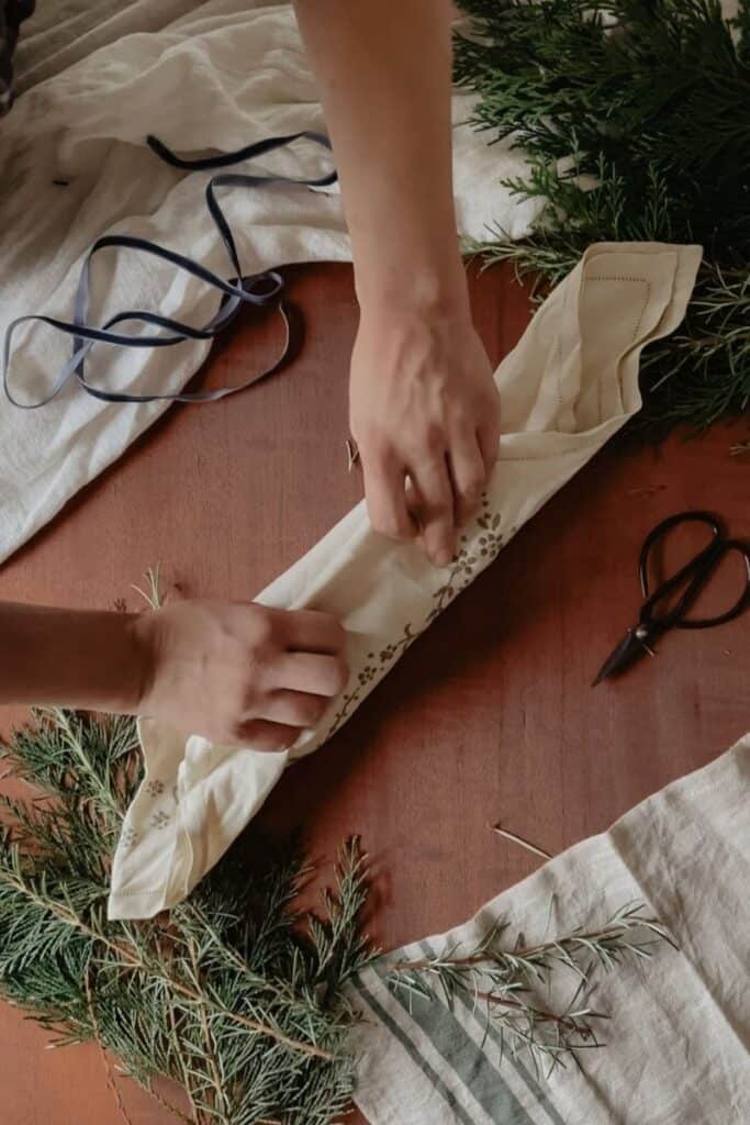 A gift being wrapped using the furoshiki method, using a vintage linen. The other corner of linen is being brought over the top of the gift