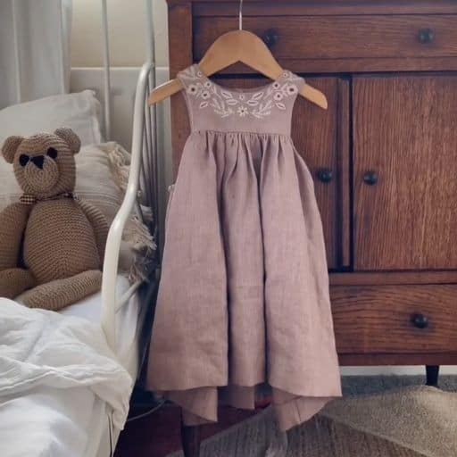 A handmade purple dress with floral embroidery hangs on an antique cupboard next to a child's bed with a knitted teddy bear on it