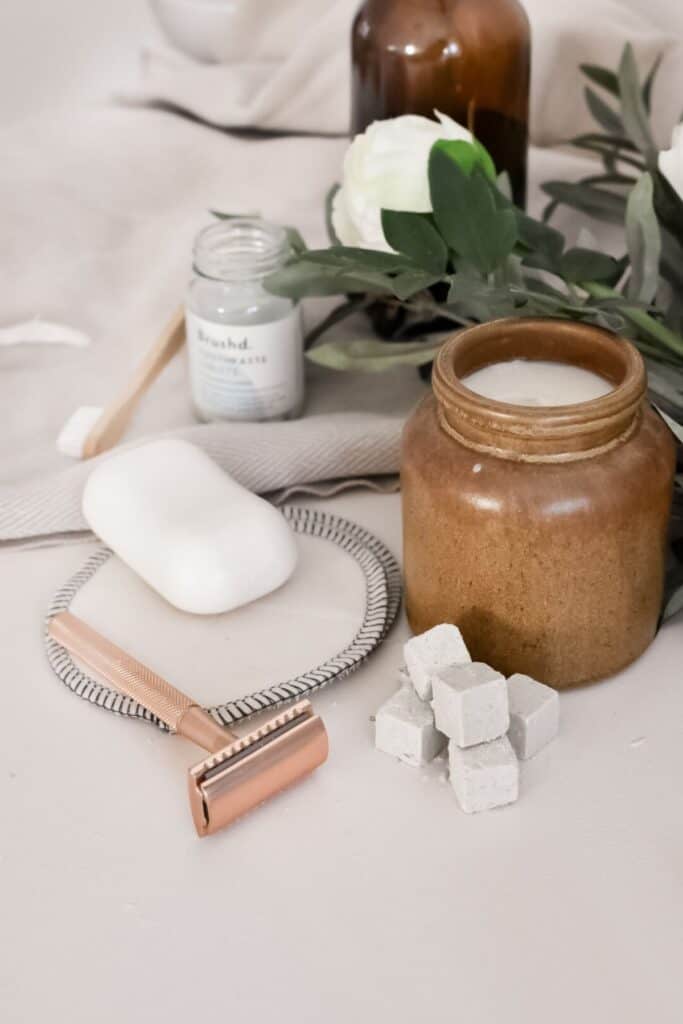 Eco-friendly bathroom products are arranged on a surface: a bar of soap on a dish, a copper-colored safety razor, a brown jar with a lid, cubes of solid material, a jar of toothpaste, and greenery with a white flower in the background.