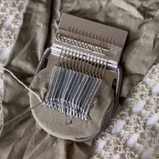 A darning loom with warp threads in blue and white and weft threads being threaded through. The threads are forming a gingham pattern.