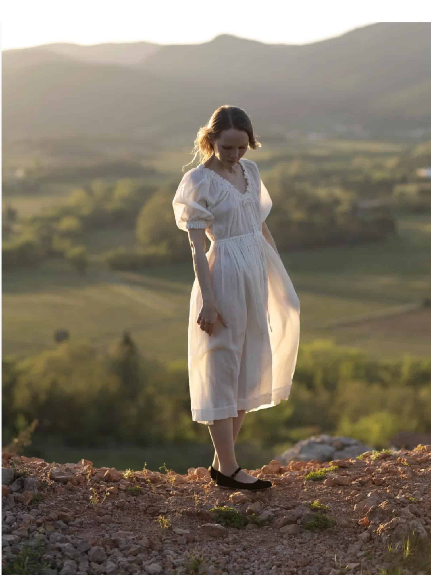 A woman in the Silversage Eleonora Dress, a romantic white dress with puffed sleeves and delicate ruffles, standing on a rocky hill at sunset, with rolling green hills in the background.