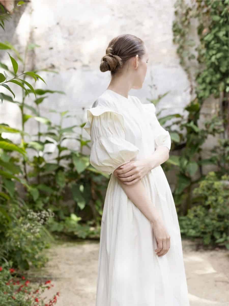 A woman in the Silversage Ophelia Dress, a flowing, vintage-inspired white gown with dramatic puffed sleeves and a cinched waist, standing in a lush, ivy-covered garden.