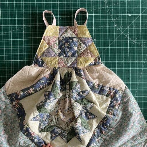 An upcycled girls pinafore made from an old floral patchwork quilt lies on top of a green cutting mat
