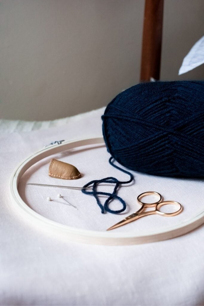 A ball of dark blue yarn, a pair of gold scissors, two sewing needles, and a thimble rest inside an embroidery hoop on a white fabric-covered surface. An embroidered abstract cushion is being crafted nearby, with a wooden chair leg partially visible in the background.