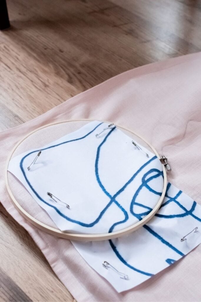 An embroidery hoop with fabric is placed on a piece of pink cloth. A white paper with a blue abstract pattern is secured with safety pins to the fabric. The setup rests on a wooden floor.