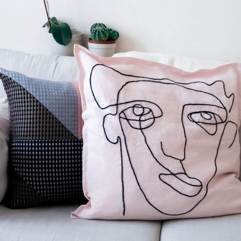 Two decorative pillows grace a white couch, one with an abstract black and gray design and the other embroidered with a pink face drawing. In the background, two small potted plants add a touch of greenery.