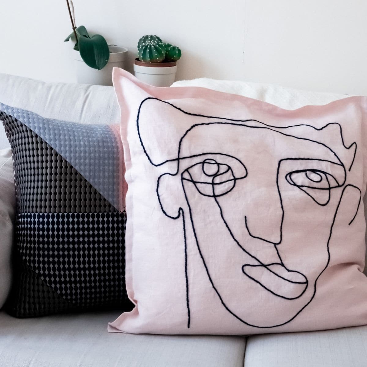 Two decorative pillows grace a white couch, one with an abstract black and gray design and the other embroidered with a pink face drawing. In the background, two small potted plants add a touch of greenery.