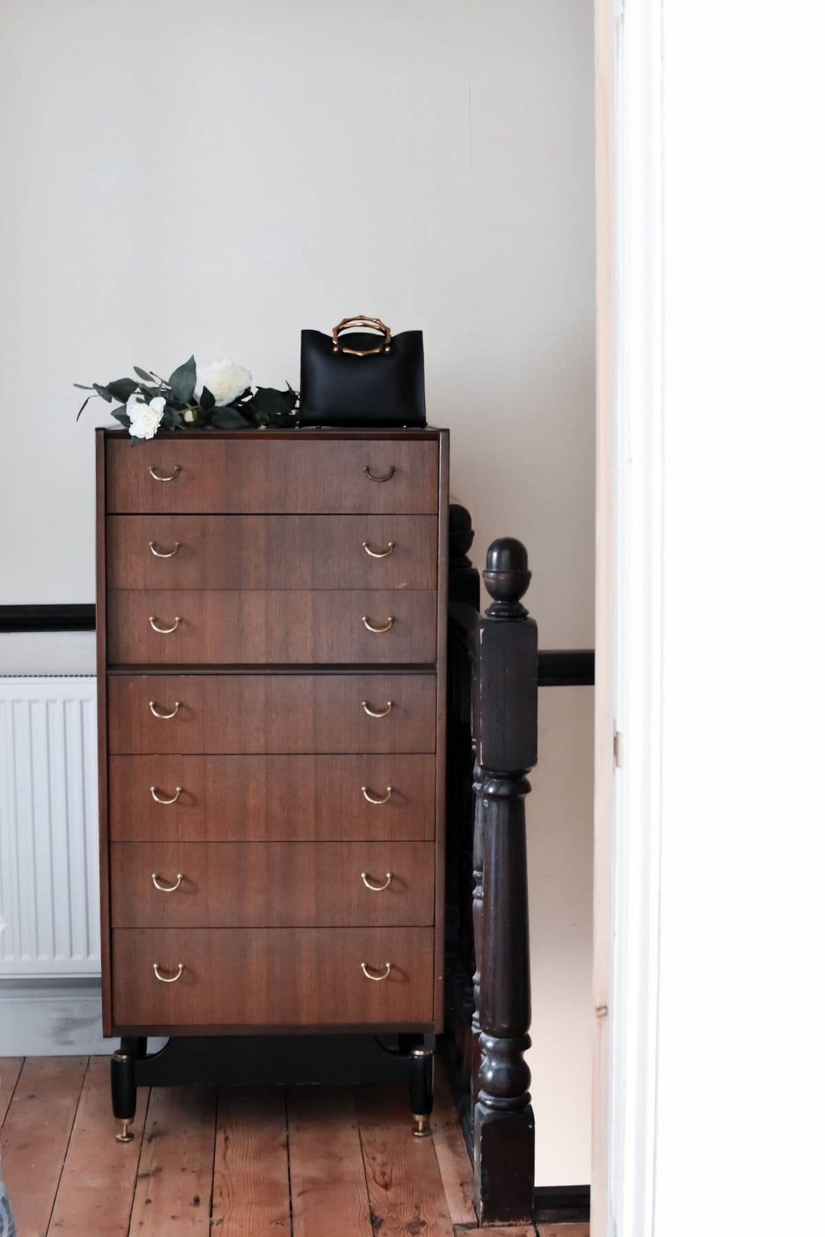 An antique bargain, this wooden dresser with five drawers and brass handles rests against a wall on the wooden floor. A black handbag and white flowers adorn the top. A portion of a radiator is visible to the left, while a dark wooden railing frames the right.