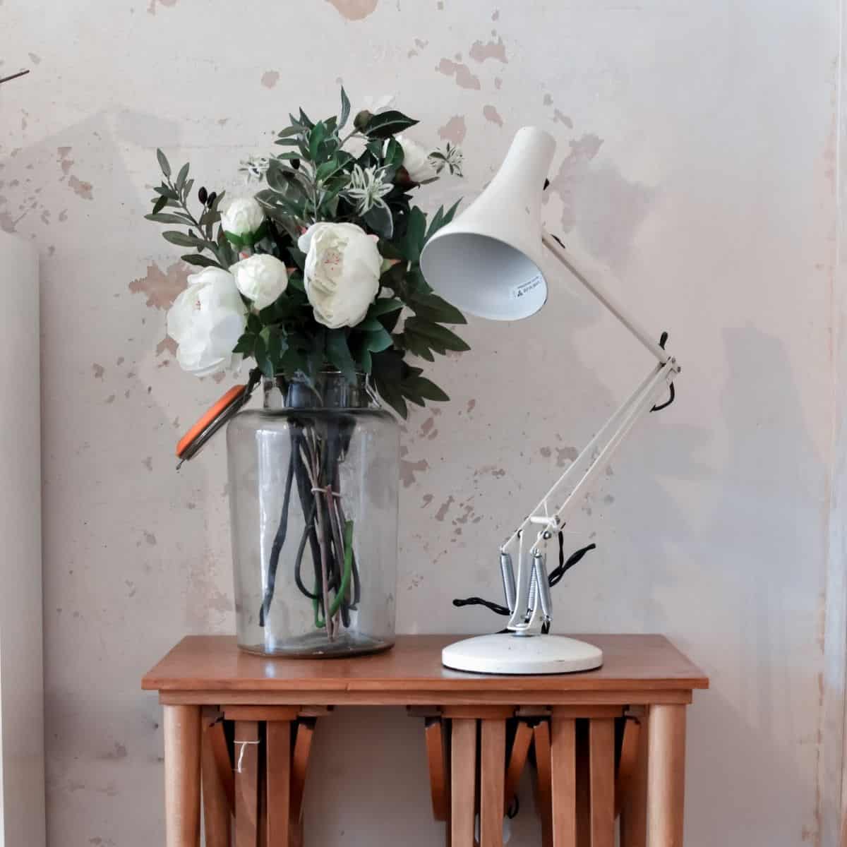 A wooden table holds a glass vase with white flowers and greenery beside a white anglepoise desk lamp, reminiscent of an antique find. The background features a textured beige wall with patches of exposed plaster, all suggesting a bargain hunters dream on eBay.