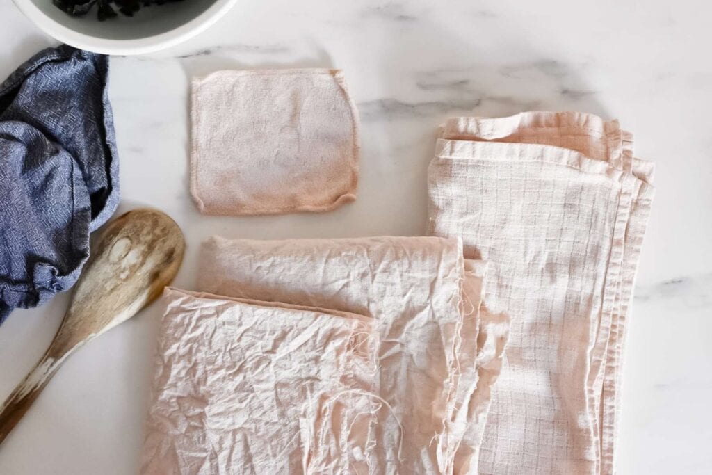A wooden spoon, crumpled cloths in shades of pink from natural avocado dye, and a white bowl with avocado skins rest on a white marble surface.