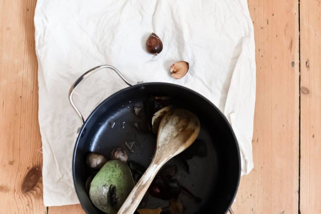 A black pot containing avocado pits and skins with a wooden spoon sits on a wooden surface, partially covered by a white cloth. A couple of avocado pits are visible next to the pot, hinting at its use for creating natural fabric dye.