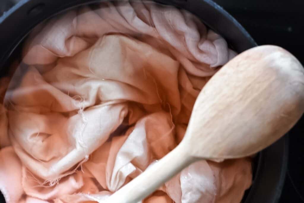 A close-up of natural fabric dye at work, featuring fabric immersed in a pot of water, tinged with shades of pink and peach. Avocado pits lend their unique hue, while a wooden spoon rests on the edge, partially submerged alongside the vibrant fabric.