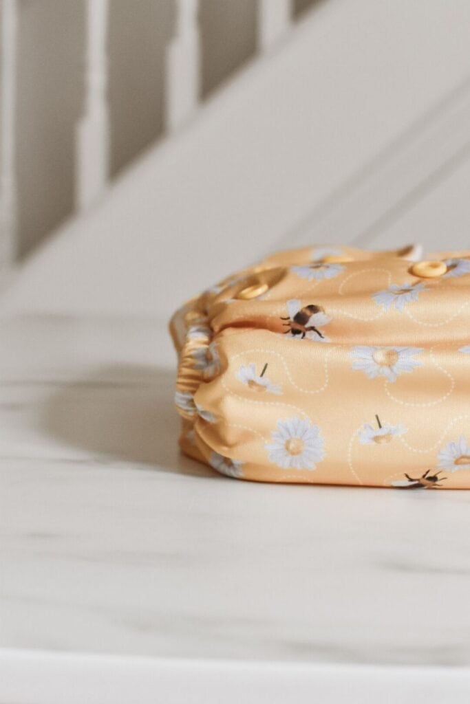 The folded yellow Baba and Boo cloth nappy, adorned with white daisy patterns and tiny insects, rests gracefully on a white marble surface. A blurred staircase in the background adds depth, reminiscent of a serene Baba + Boo review setting.