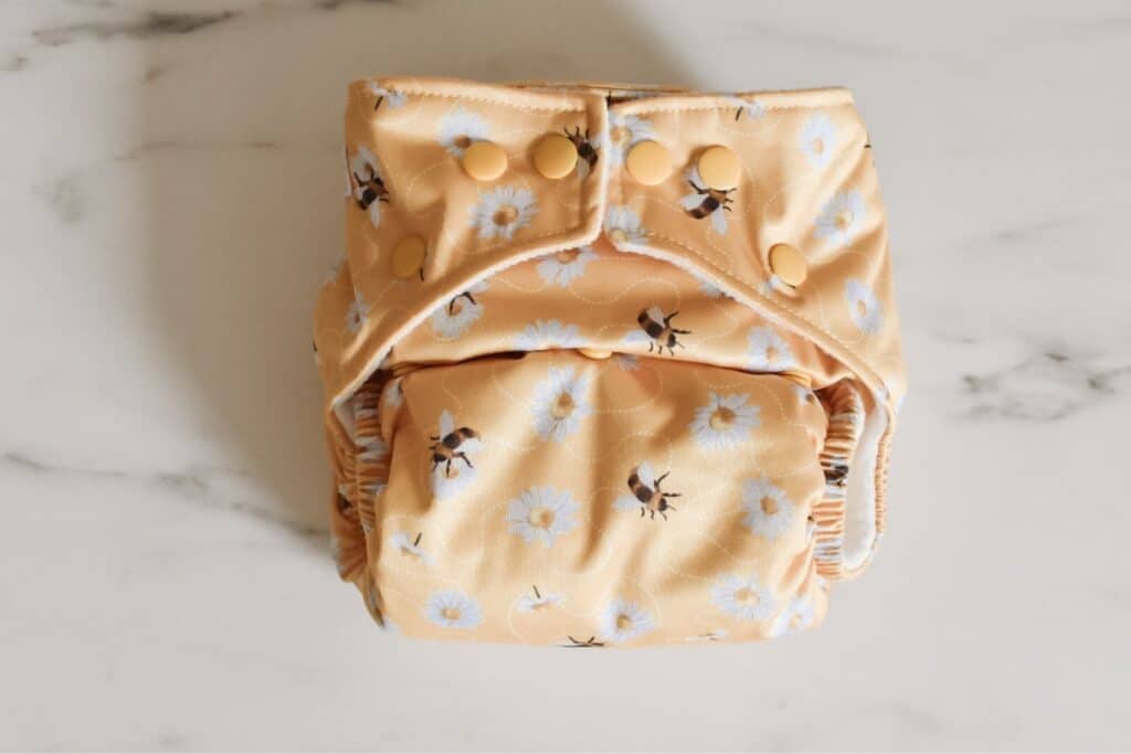 The Baba + Boo reusable cloth nappy, adorned with a yellow floral and bee design, rests on a light marble surface. Featuring snap buttons for adjustable sizing, this versatile one-size nappy promises both style and practicality.