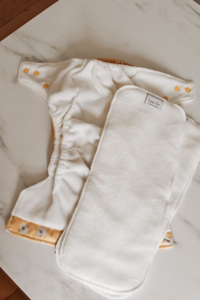Cloth nappy and insert set from Baba + Boo on a marble surface. The nappy features yellow buttons and floral details, while the plain white insert emphasizes an eco-friendly, reusable design. These one-size nappies are perfect for sustainable baby care.