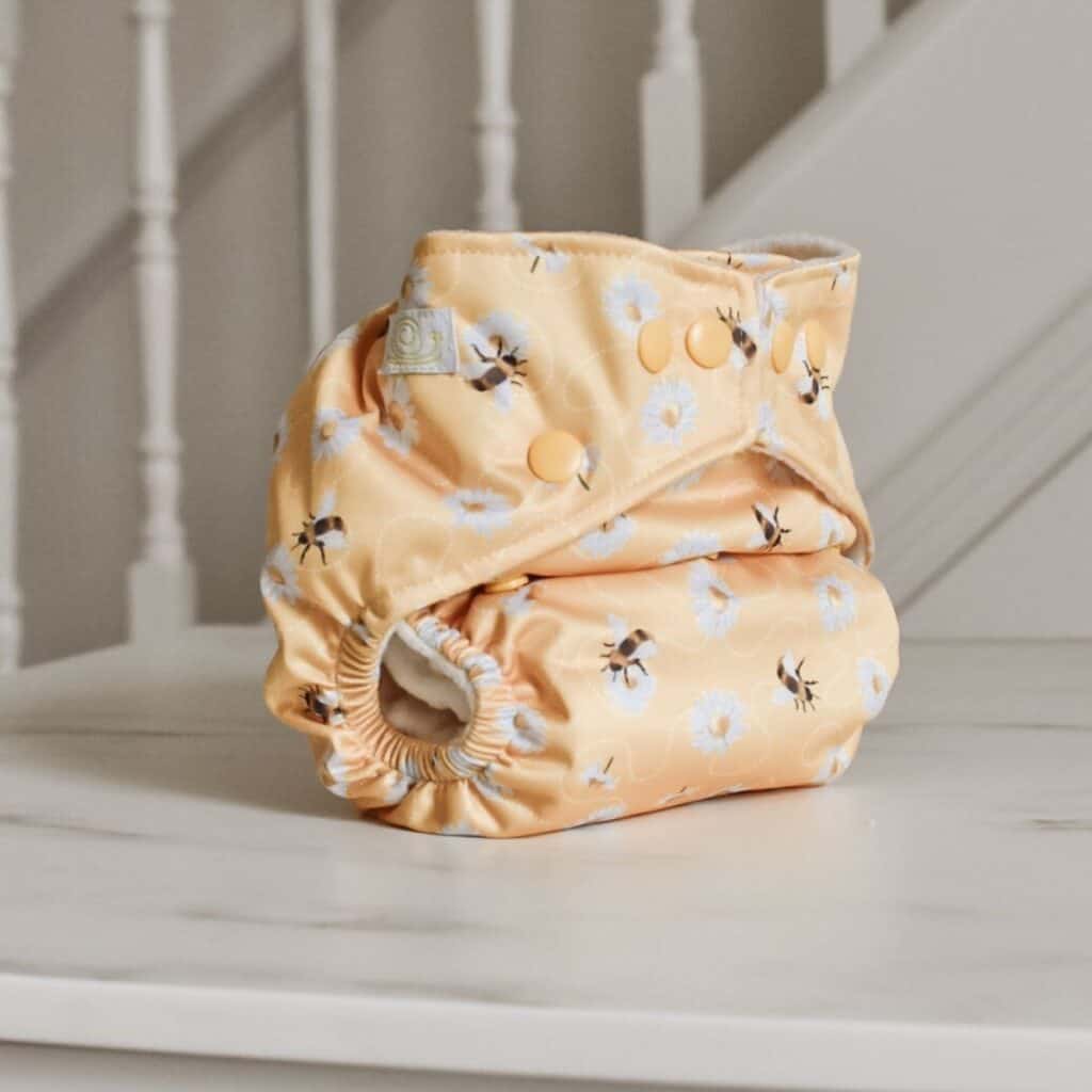 A cloth nappy by Baba + Boo, with a pastel yellow background featuring white flowers and bumblebee patterns, sits folded on a white marble surface. In the background, there is a white staircase railing. 