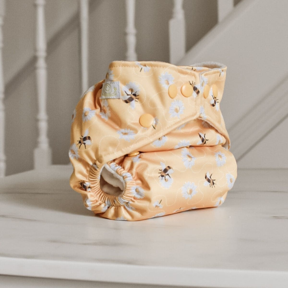 A cloth diaper by Baba + Boo, with a pastel yellow background featuring white flowers and bumblebee patterns, sits folded on a white marble surface. In the background, there is a white staircase railing. This one-size nappy combines charm and function effortlessly.