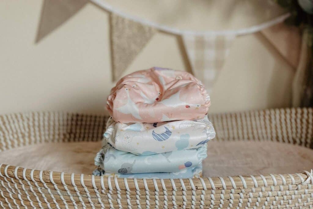 A stack of three colorful, reusable cloth nappies from Bambino Mio sits in a woven basket. The top nappy is pink with a goose design, while the others underneath are white and light blue with various playful patterns. In the background, there is a fabric bunting.