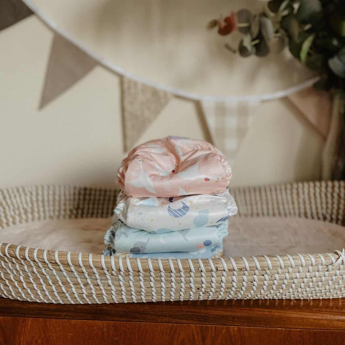 A stack of three pastel-colored cloth nappies sits on a woven changing table. The Tots Bots diapers boast playful patterns in pink, blue, and white hues. In the background, decorative hanging flags and a small plant enhance the charming scene.
