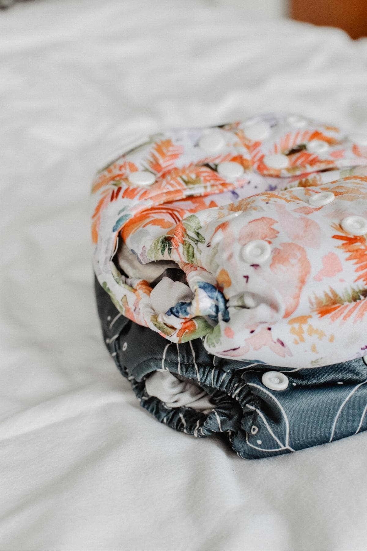 Two cloth nappies are stacked on a white bedspread. The top nappy, a Bare & Boho V3 one-size nappy, features a colorful floral pattern, while the bottom one, a V2, has a dark background with white lines and shapes.