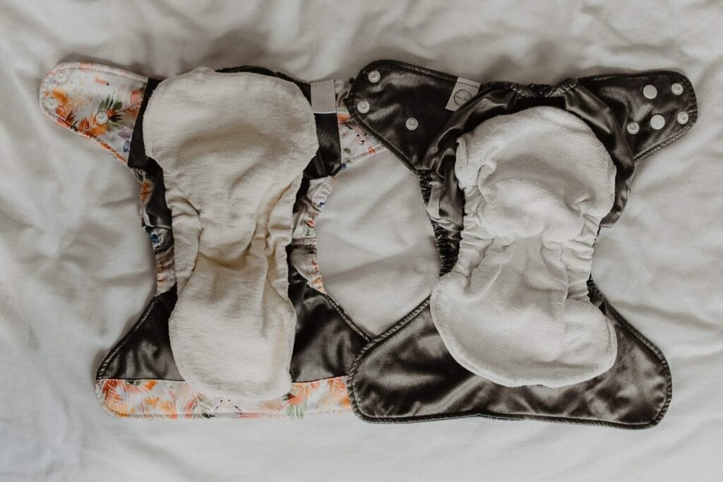 Two reusable Bare & Boho cloth nappies with inserts lay open on a white surface. The V3 nappy on the left features a floral pattern, while the one on the right is solid gray. Both have white absorbent pads in the center, promising style and practicality for your little ones needs. They are similar, except that the V3 nappy has a slightly larger absorbent insert and longer tabs.