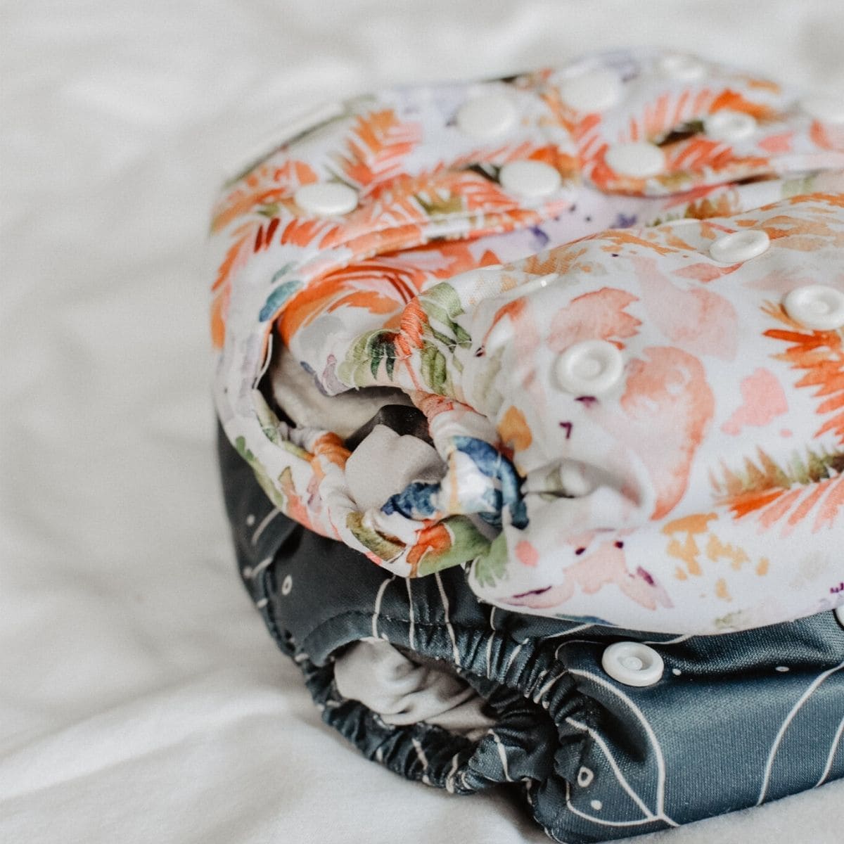 Two cloth nappies, both one sizes nappies from Bare & Boho, stacked on a white surface. The top nappy boasts a colorful floral pattern with white snaps, while the bottom showcases a darker design with matching snaps.