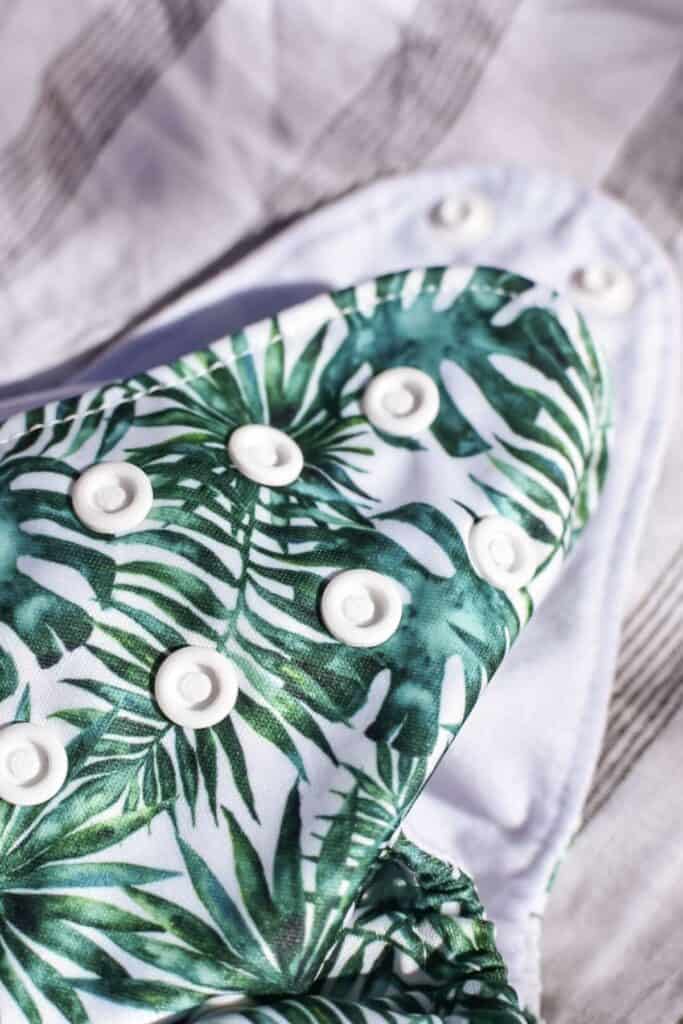 Close-up of a Bear Bott All-in-Two Nappy featuring a tropical leaf pattern and white snap buttons. The background reveals soft, light fabric with gray stripes, perfect for ensuring comfort.
