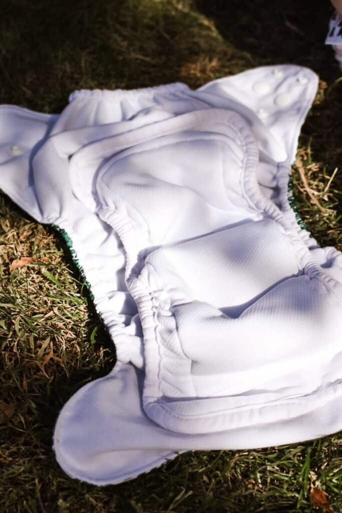 A Bear Bott All-in-Two Nappy with white fabric and button snaps lies open on grass, revealing its padded interior and elastic edges.
