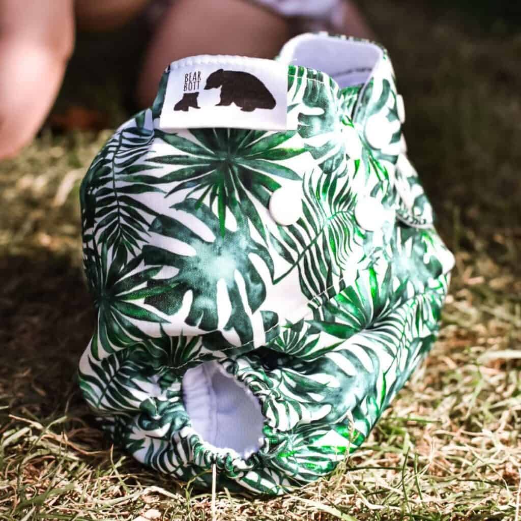 The Bear Bott All-in-Two Nappy boasts a tropical leaf print design with convenient snap closures, resting on lush grass. A charming bear silhouette adorns the label.