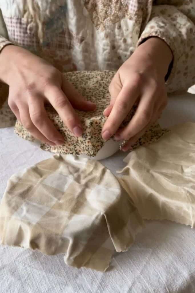 Hands wrapping a bowl with DIY beeswax wraps. Two other bowls are wrapped in beeswax wraps nearby. The person wears neutral-toned clothing.