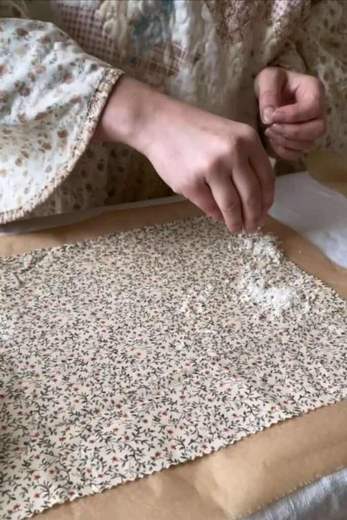 Wearing a floral-patterned garment, a person demonstrates how to DIY beeswax wraps by sprinkling grated beeswax onto fabric adorned with small florals. The fabric rests on brown parchment paper, ready to be ironed.