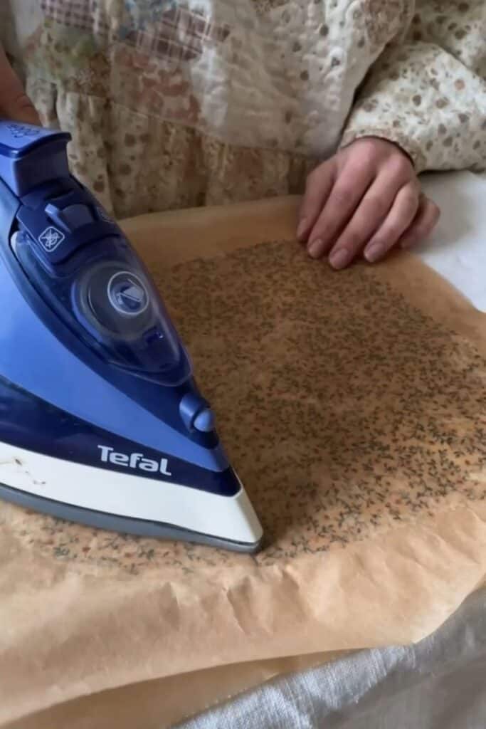 A person skillfully irons fabric with a blue Tefal iron, melting the beeswax onto the fabric. The floral-patterned fabric is protected under parchment paper. 
