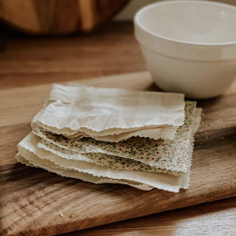 A stack of DIY beeswax wraps with floral patterns and gingham rests on a wooden board beside a white ceramic bowl on a wooden surface.