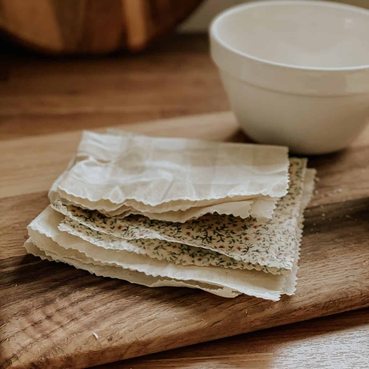 A stack of DIY beeswax wraps with floral patterns and gingham rests on a wooden board beside a white ceramic bowl on a wooden surface.