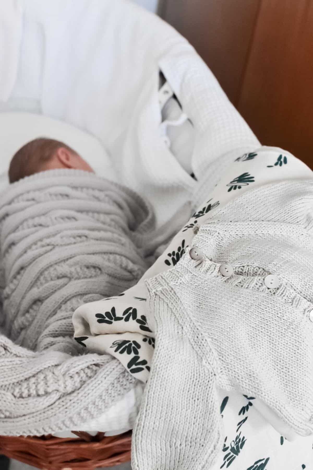 A baby, after a positive second birth, is now peacefully sleeping in a wicker bassinet, swaddled in a gray knit blanket. Nearby, a gray knitted sweater and a white cloth with green leaf patterns are draped over the side.