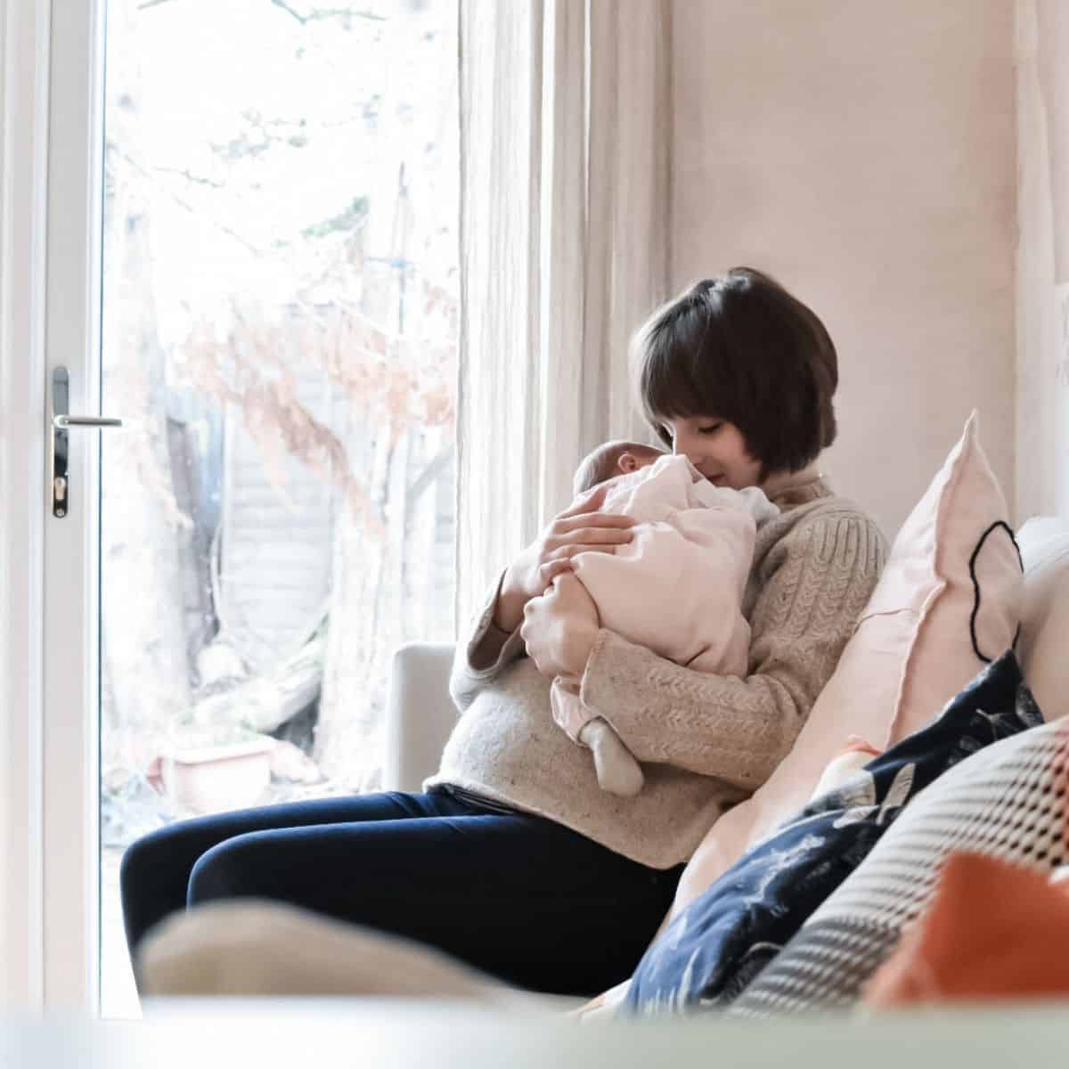A person with short hair sits on a couch, cradling a baby wrapped in a soft blanket after their positive second birth. Natural light pours in through the window behind them, illuminating the cozy setting with pastel-colored cushions.
