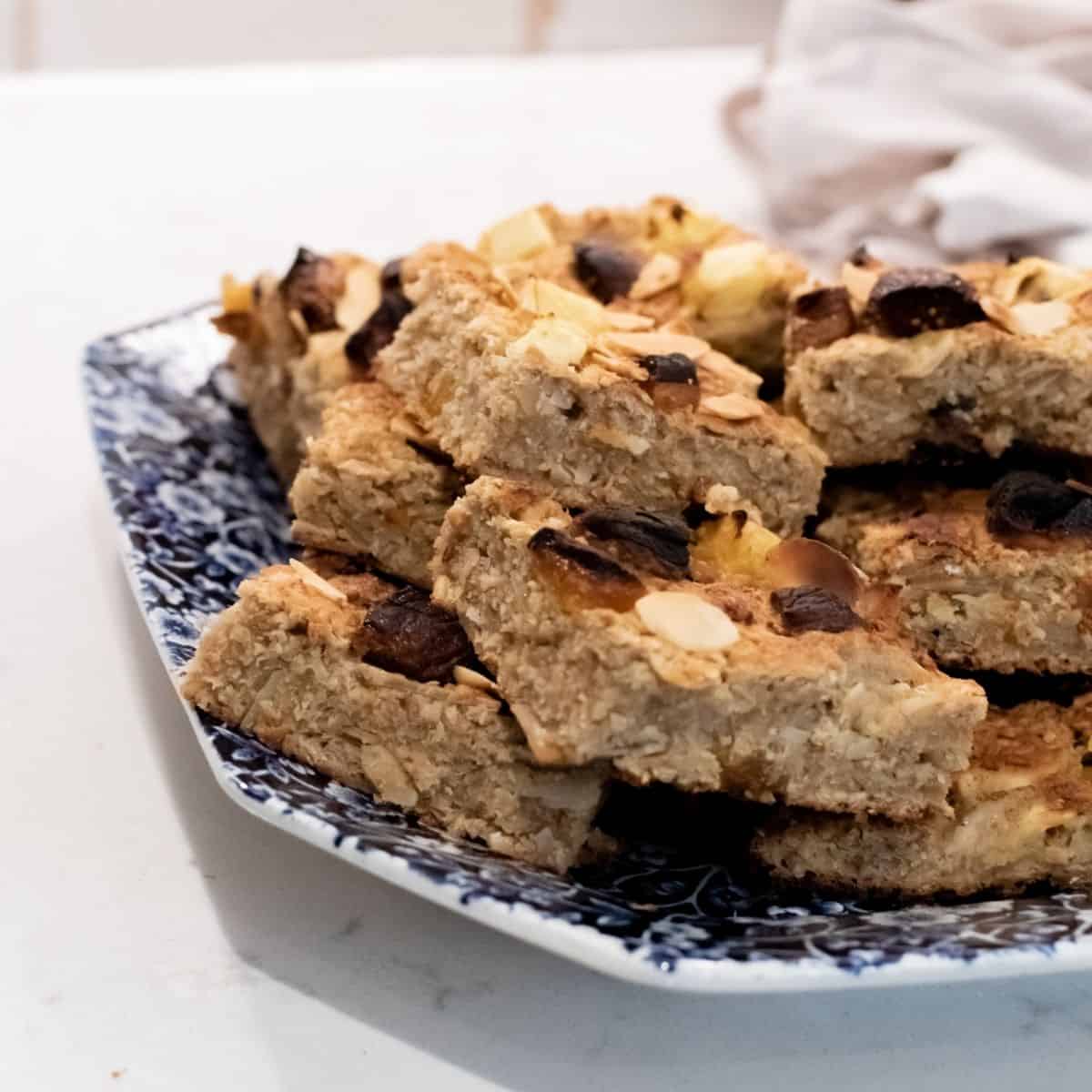A delicious recipe for homemade fruity oat bars stacked with nuts and dried fruits, served on a blue and white patterned plate. The breakfast bars are golden brown, with visible almond slices and raisins, placed on a white countertop.