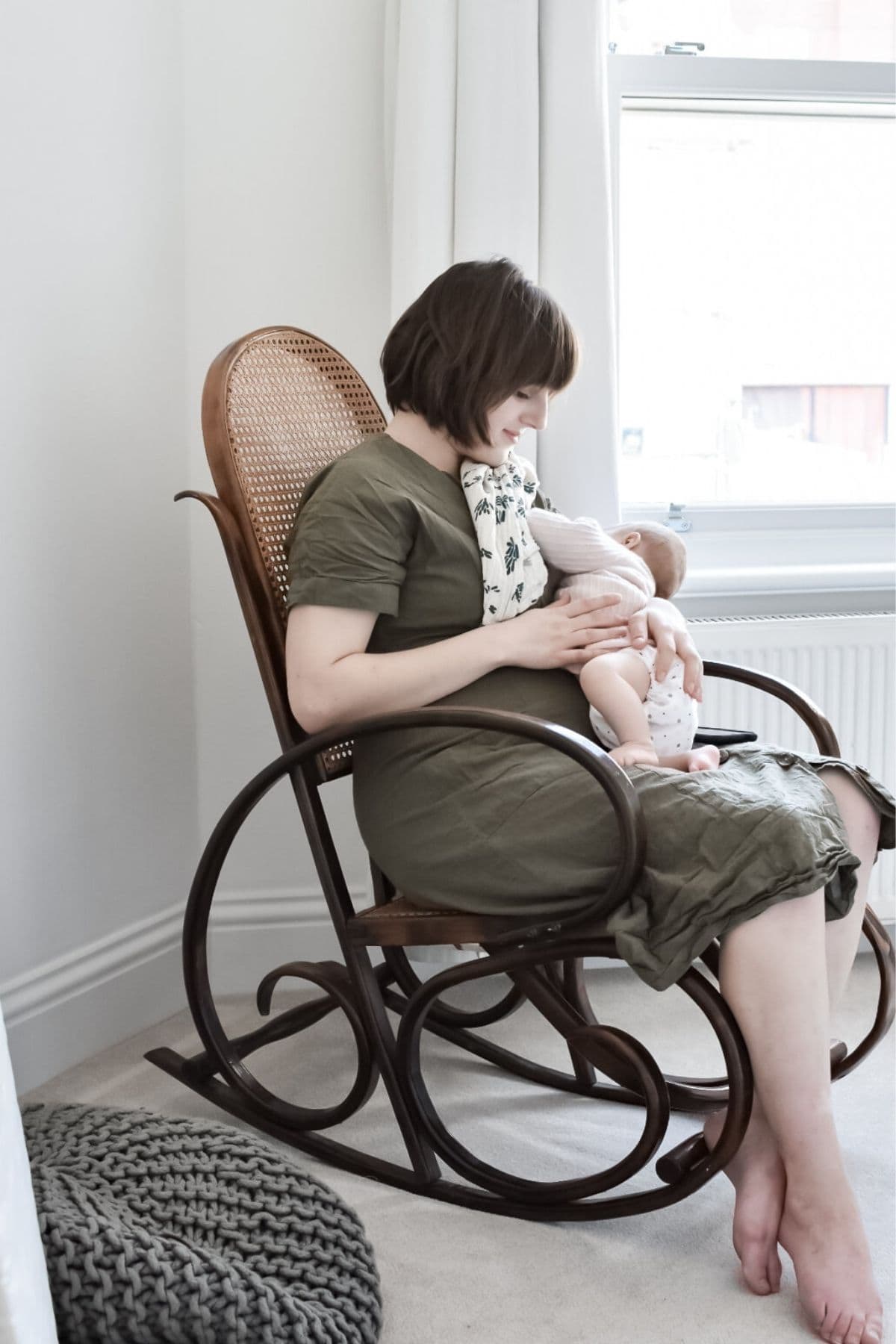A woman in a green dress sits in a wooden rocking chair, cradling her baby in an intimate moment of motherhood. The room is softly lit, with a window and a crocheted cushion on the floor. She gazes lovingly at the baby she is breastfeeding.